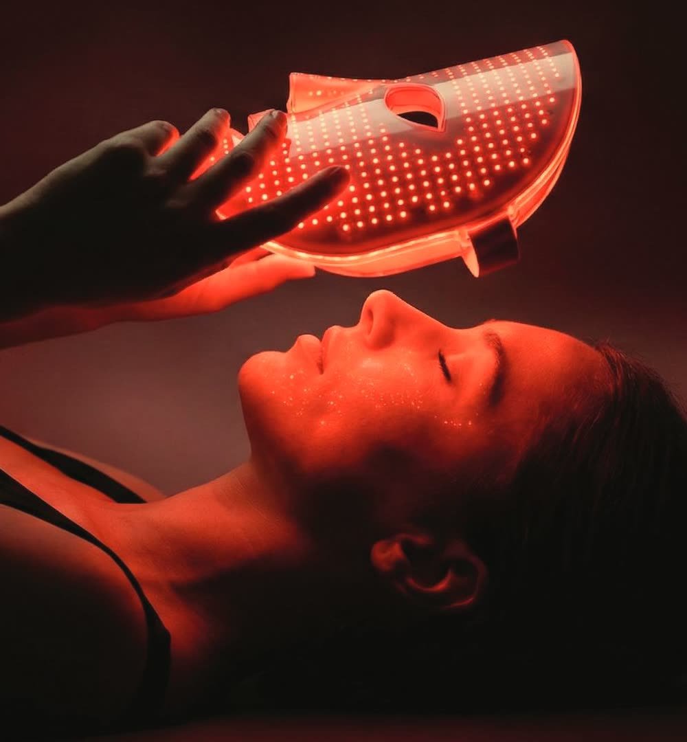 A woman lying back under a glowing red light therapy mask.